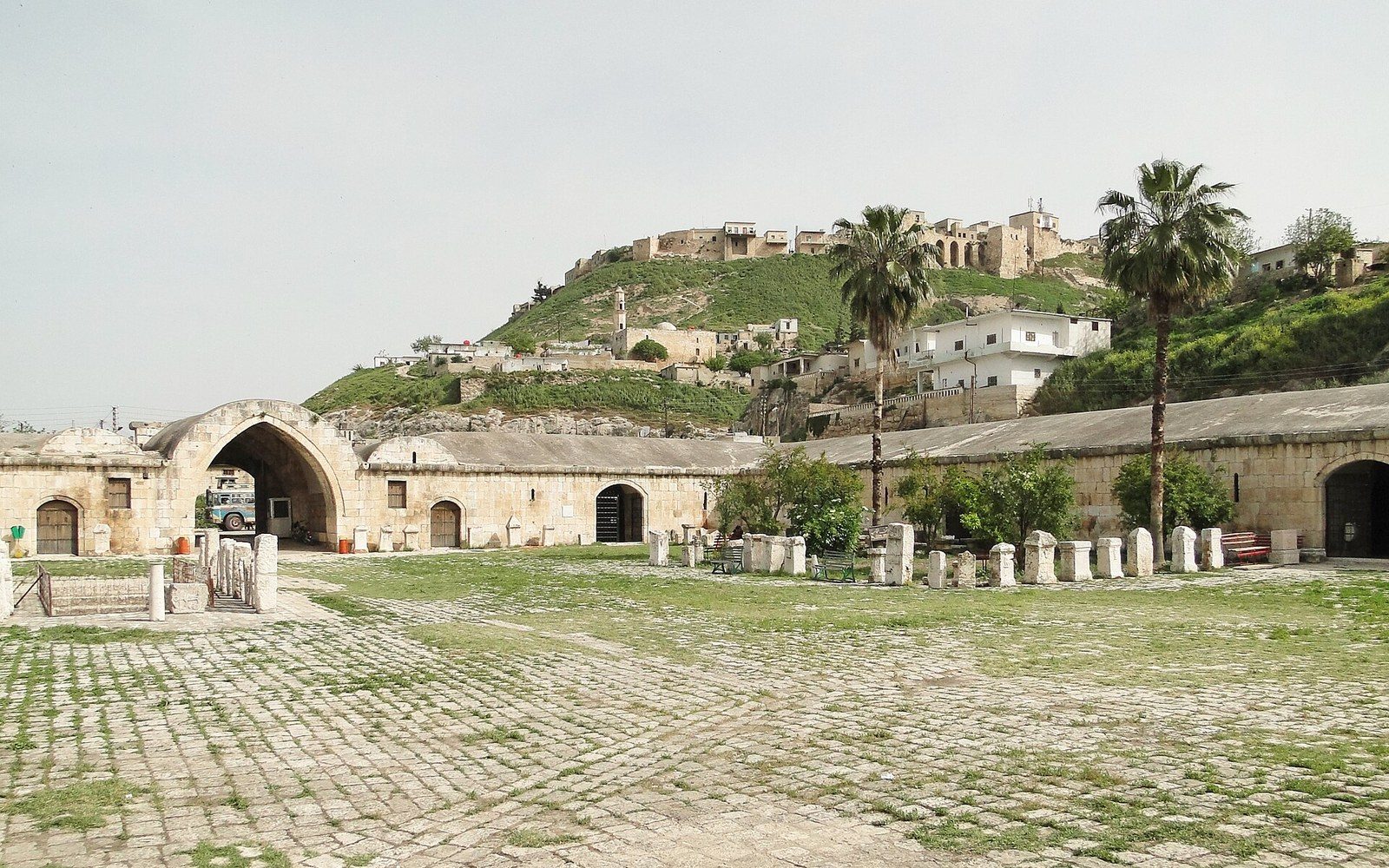 Stone caravanserai courtyard with arched arcades below a hilltop citadel and palm trees