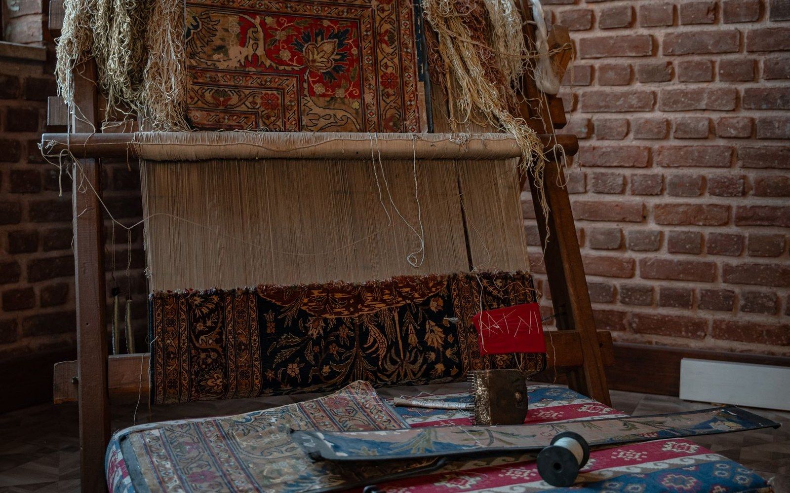 A traditional wooden loom holds a partially woven patterned carpet against a brick wall