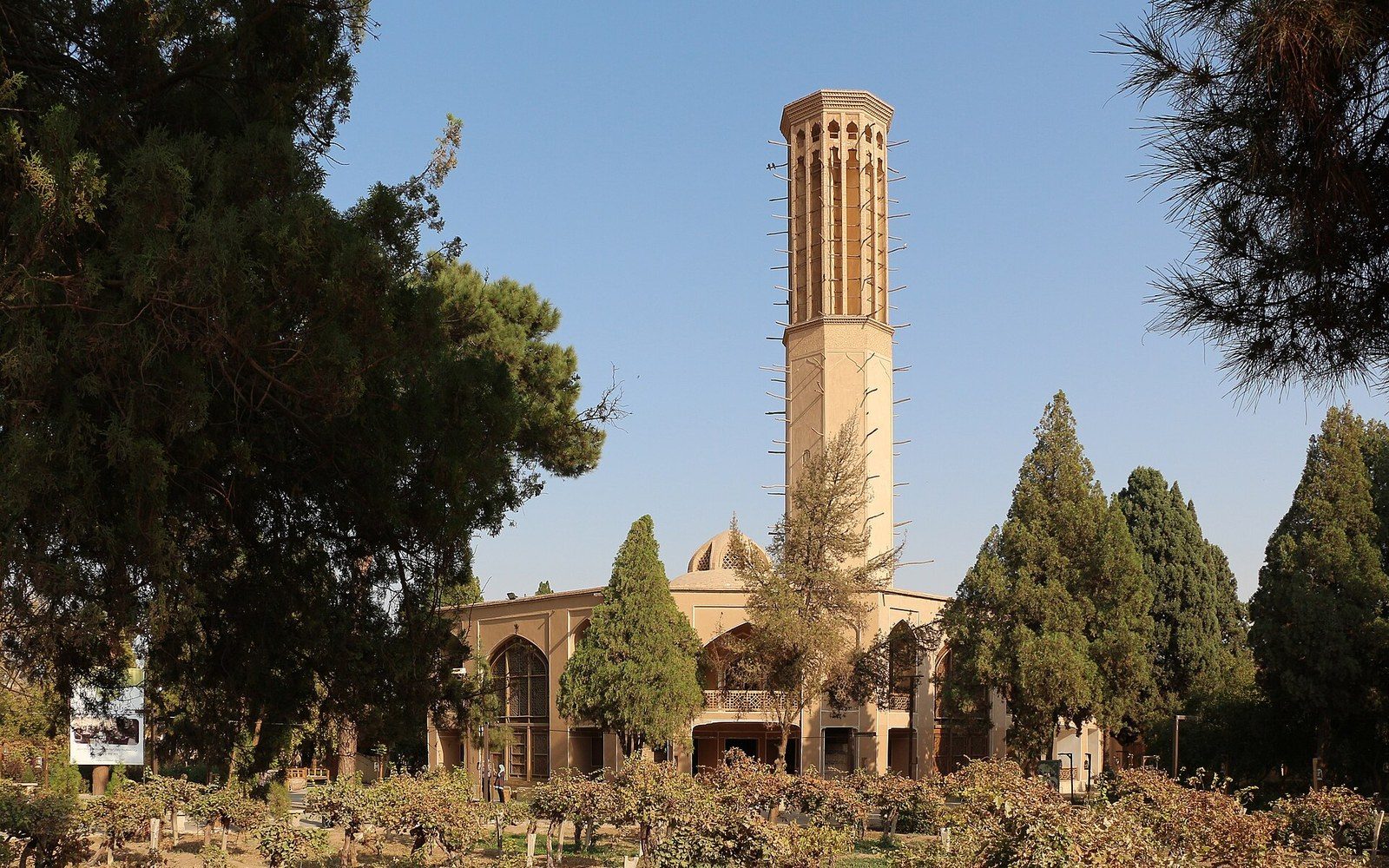 Tall traditional windcatcher tower rising above a domed pavilion surrounded by garden trees