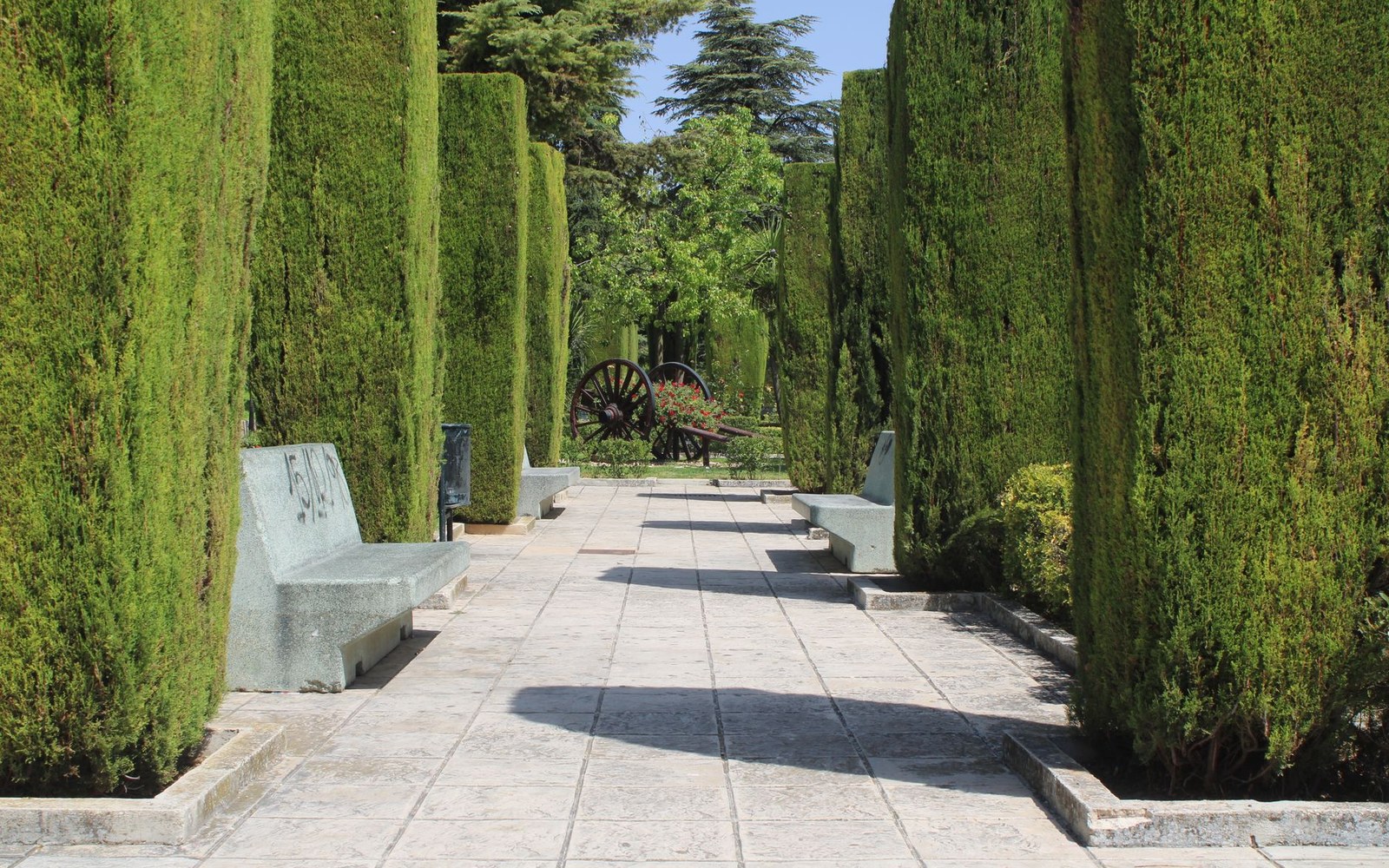 Stone benches line a hedge-bordered garden path leading to an old wooden cart