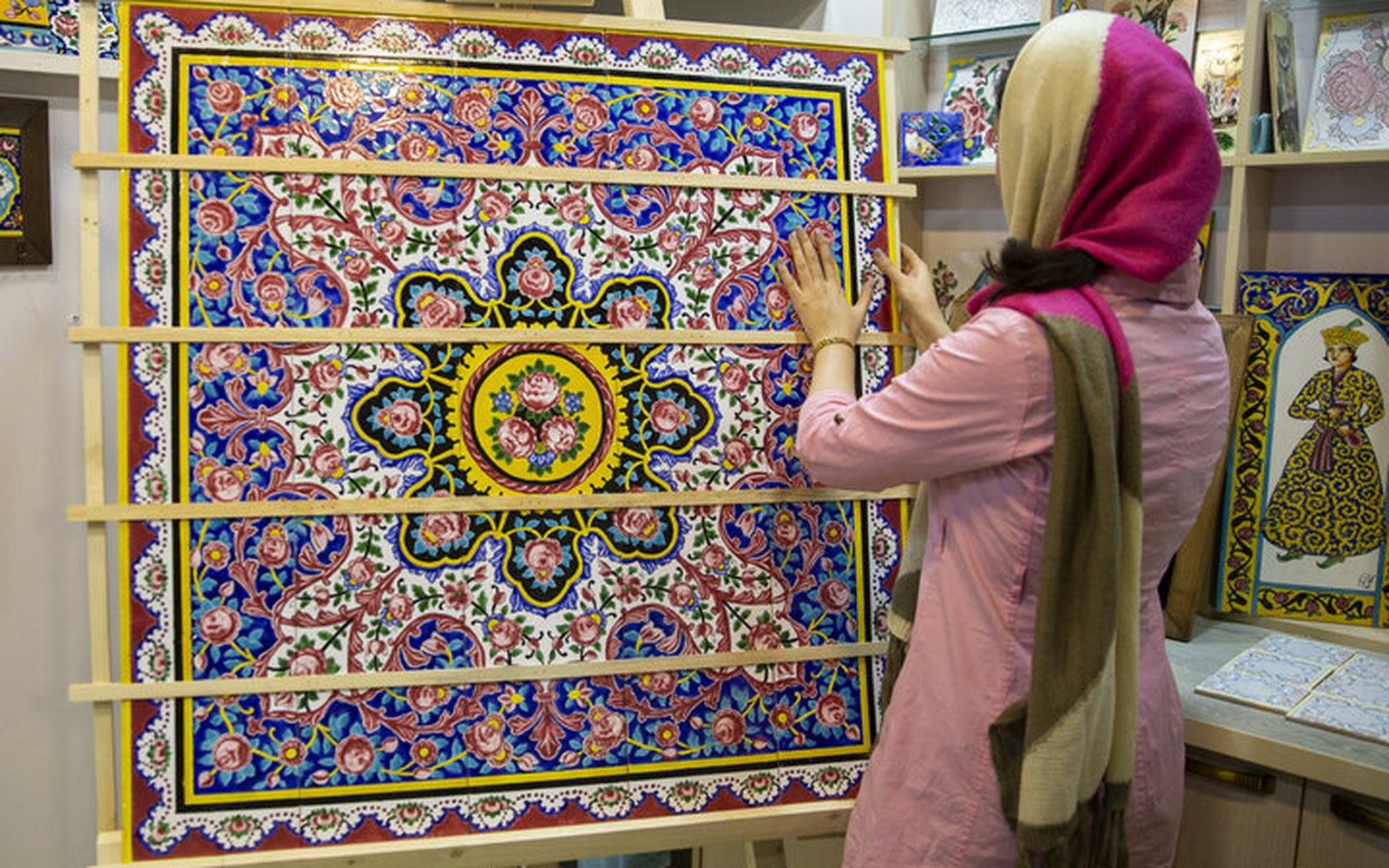 A woman examines a large tile panel featuring intricate floral and paisley patterns in a workshop