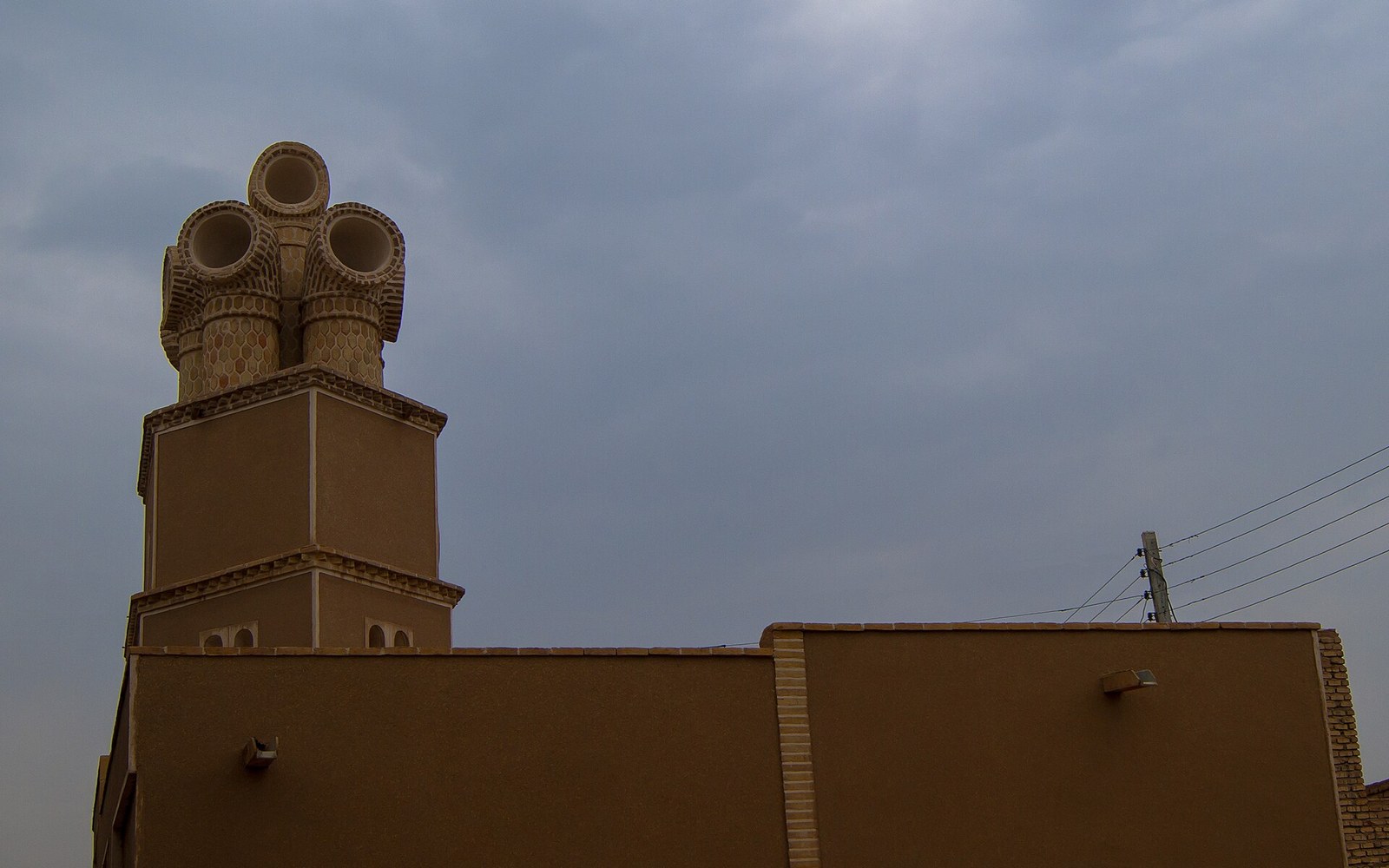 Cylindrical windcatcher towers rise above a flat-roofed adobe building under a cloudy sky