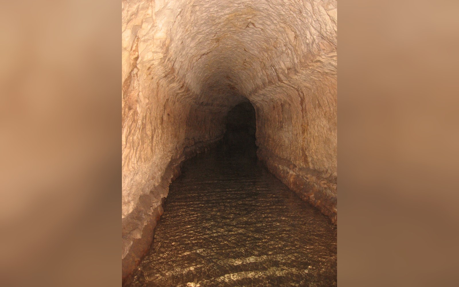 A narrow rock-cut tunnel with shallow flowing water leading into darkness