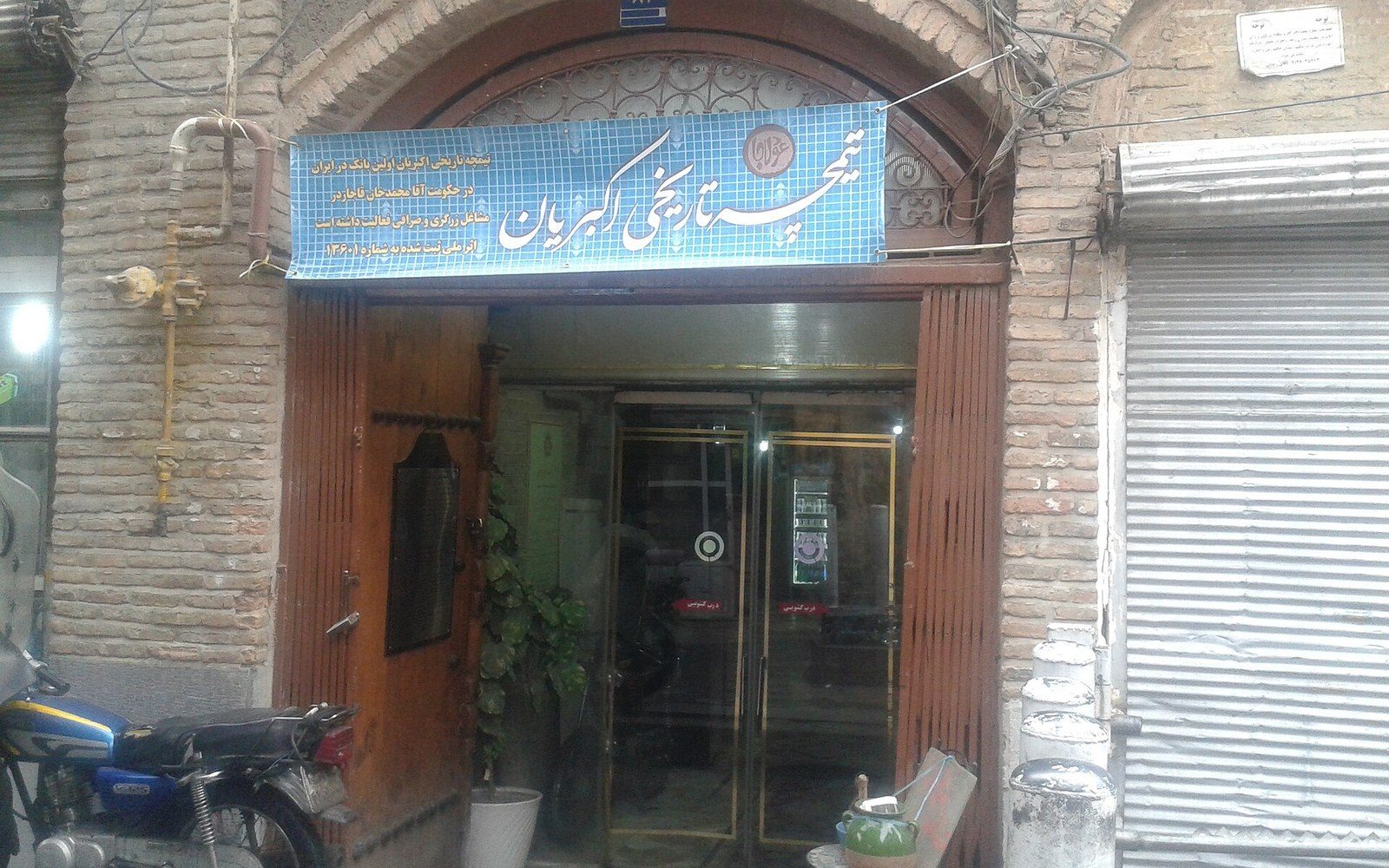 Brick building entrance with glass doors, a Persian signboard, and a parked motorcycle