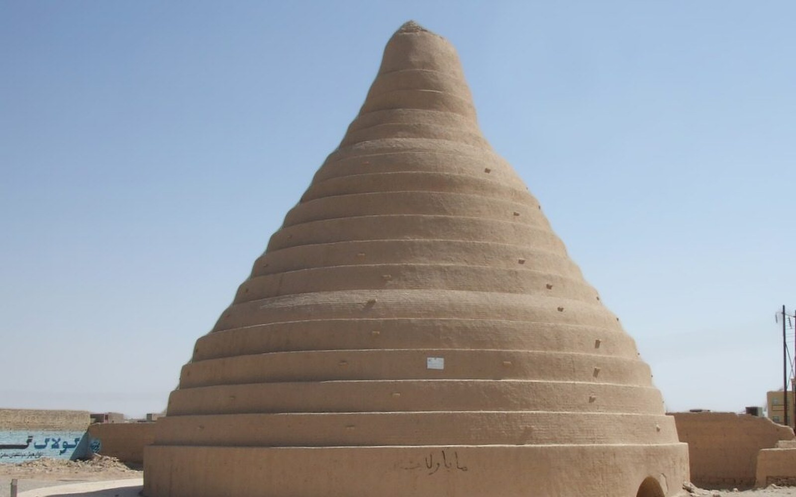 A conical mud-brick yakhchal with ringed exterior walls stands against a clear sky